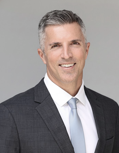 Smiling man in suit & tie. Silver hair, light skin, gray background. Professional headshot.