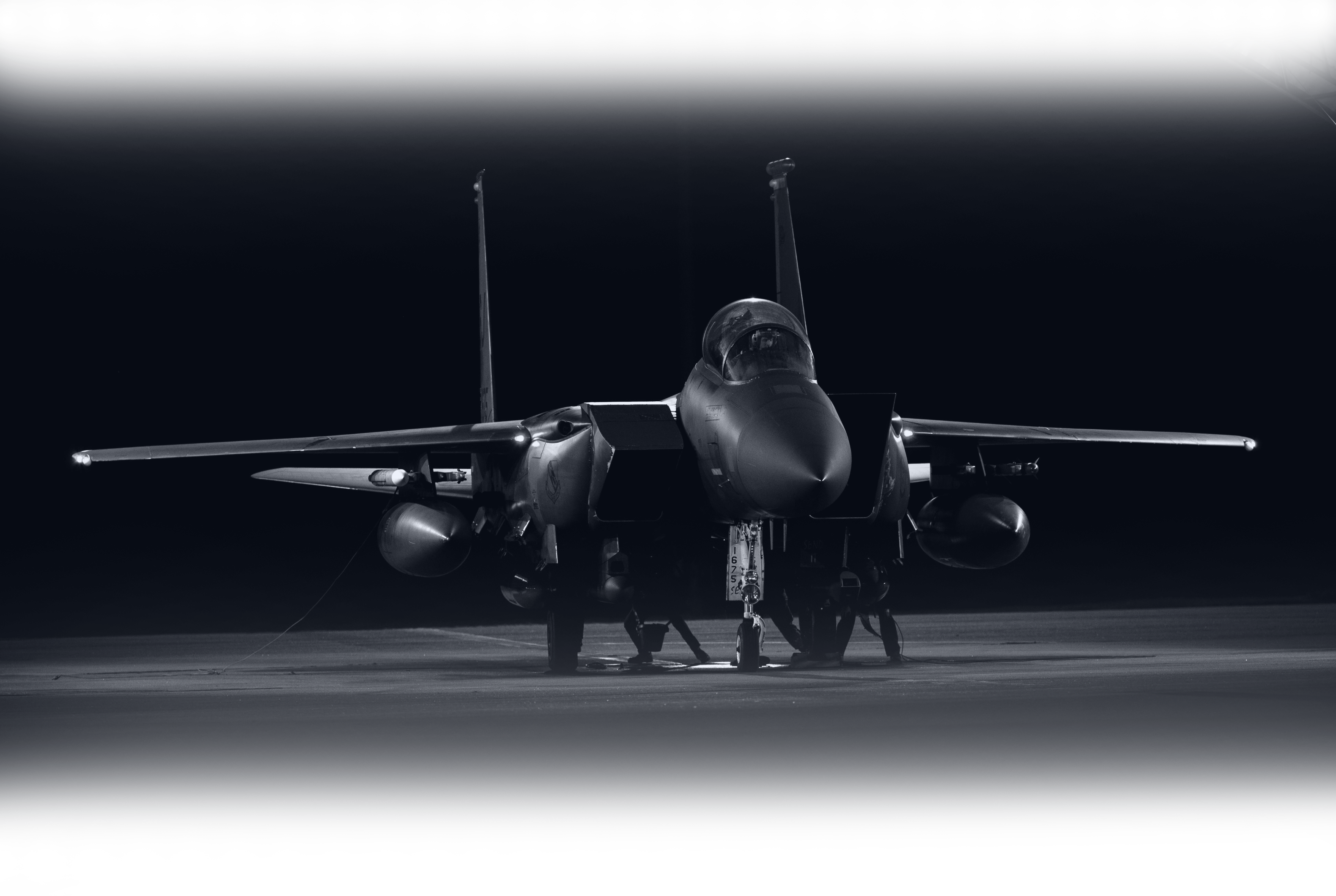 Monochrome shot of an F-15 Eagle fighter jet on the ground at night, lit by spotlights.