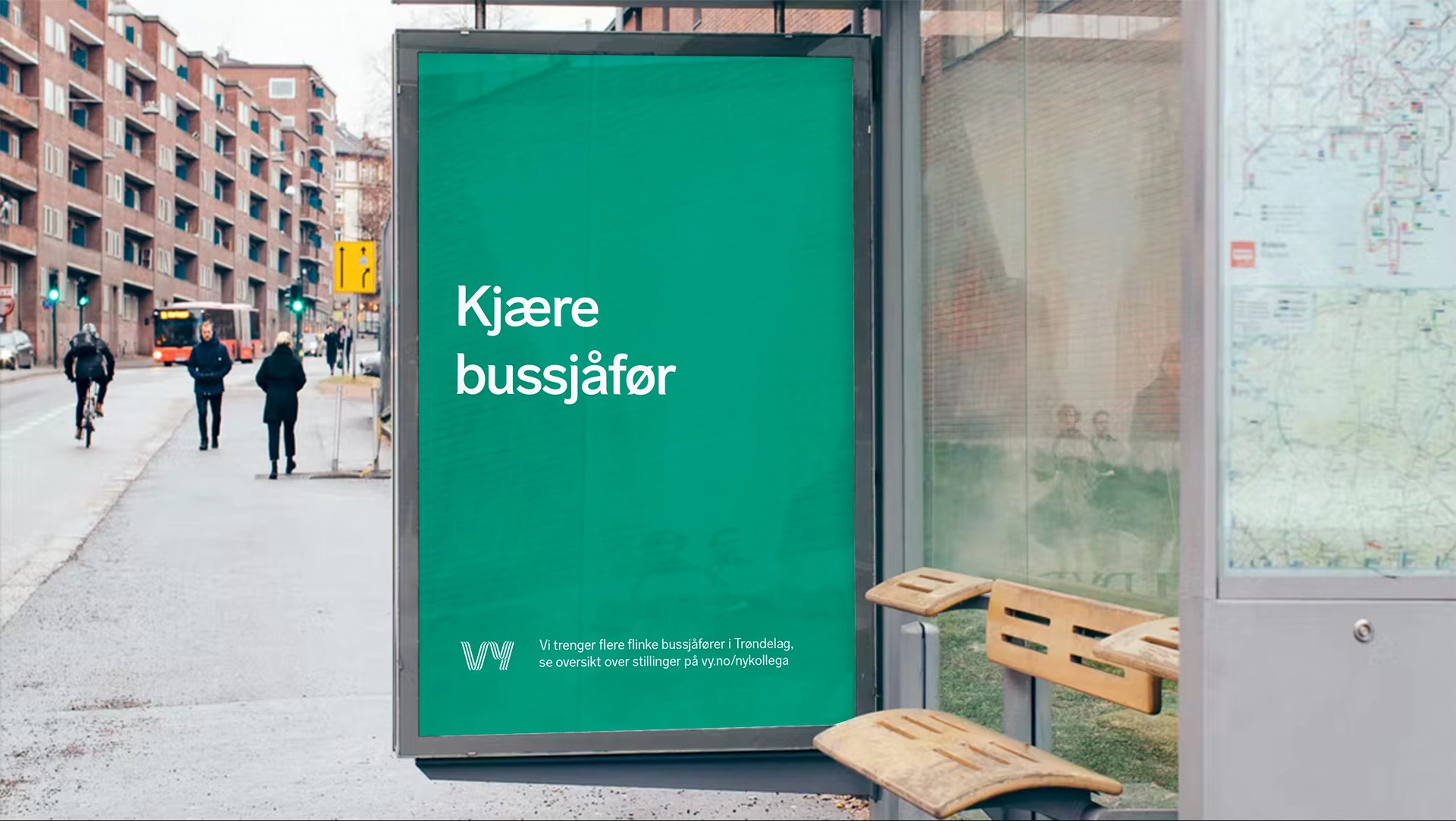 Grønn plakat i et busskur med teksten «Kjære bussjåfør», med en Vy-buss i gata bak
