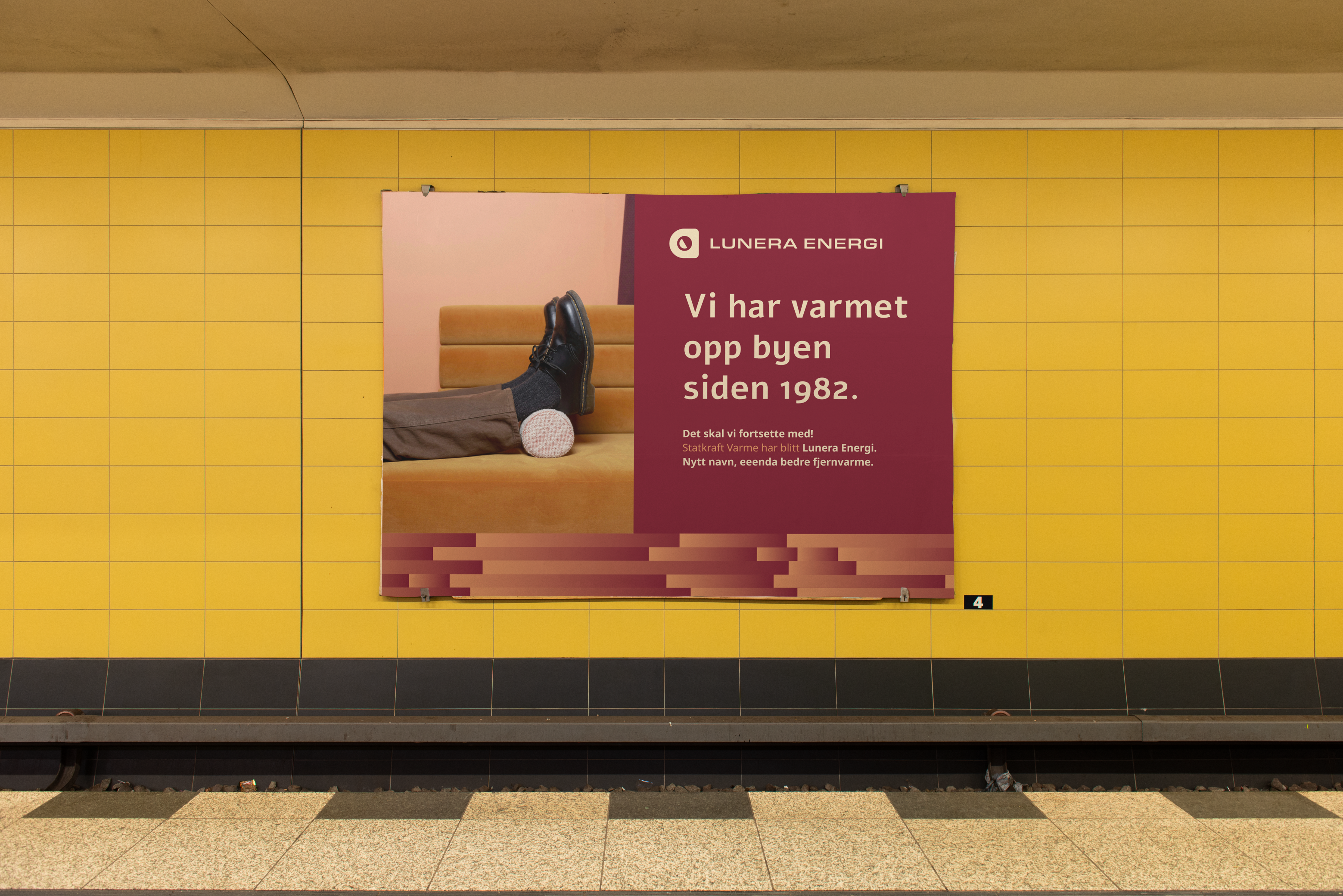 Plakat på en gul flisvegg på en stasjon: «Vi har varmet opp byen siden 1982», med et par ben strukket ut på en oransje benk.