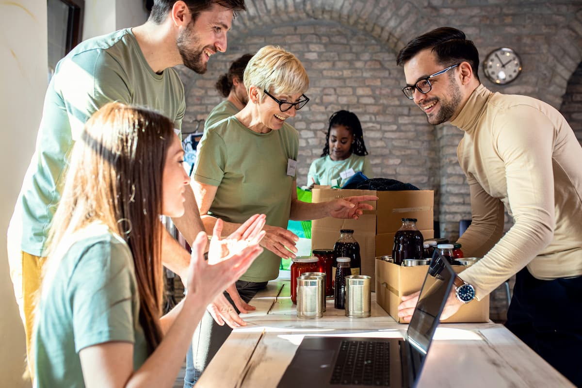 A group of people standing around a table collaborate. A woman speaks from behind a laptop, a man carries a box full of bottles.