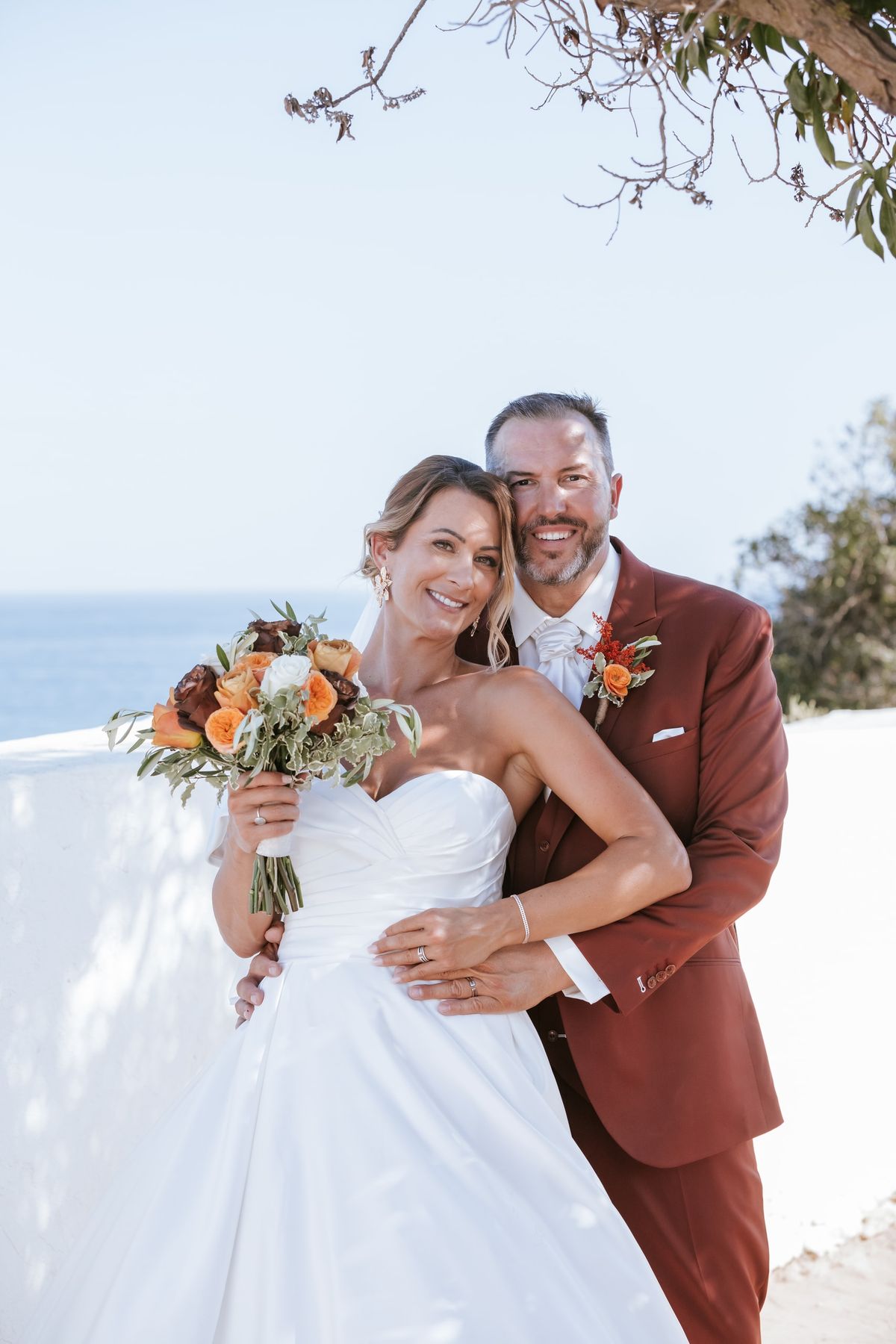 Bride and groom on a clifftop terrace above the sea, her bouquet in warm autumn tones