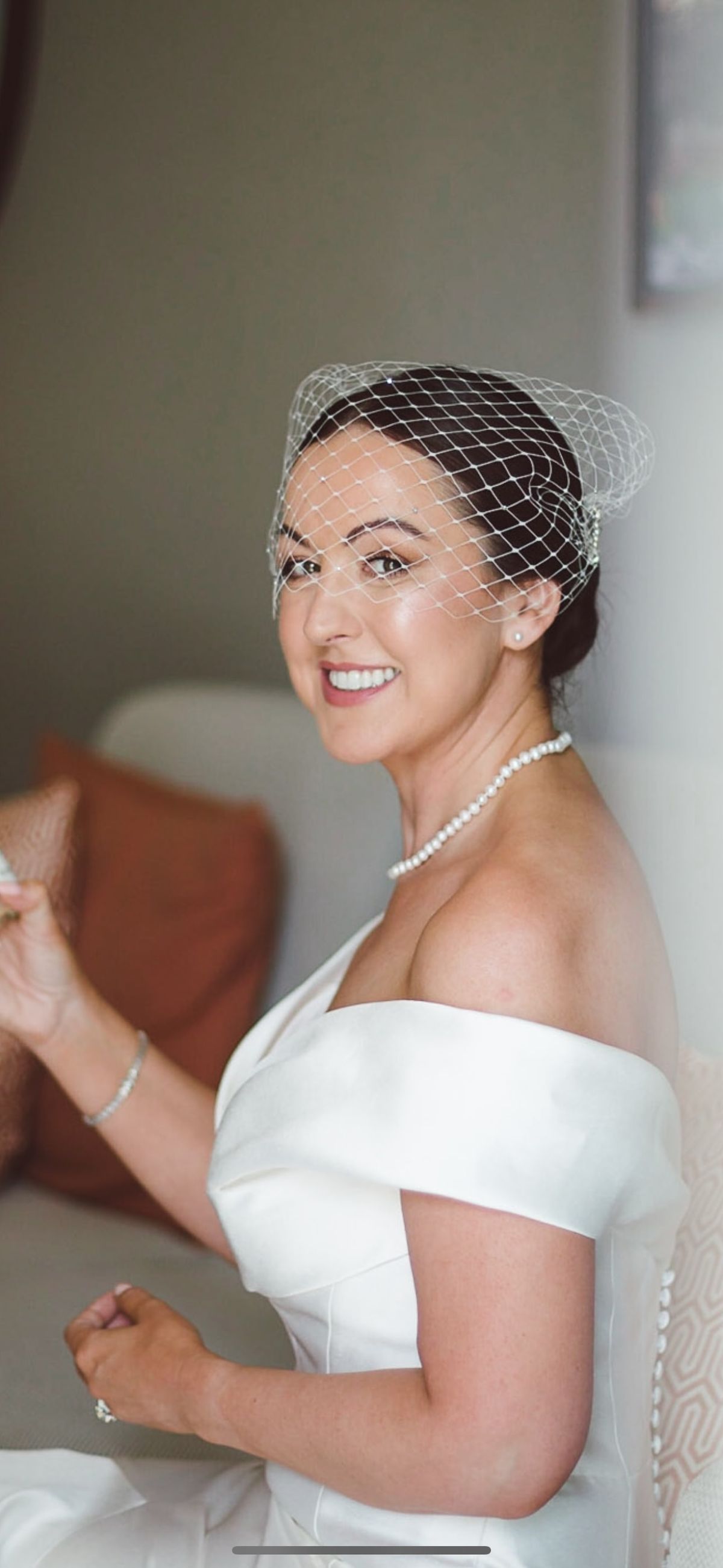 Bride in a birdcage veil and off-the-shoulder gown, smiling, with luminous skin and a pearl necklace