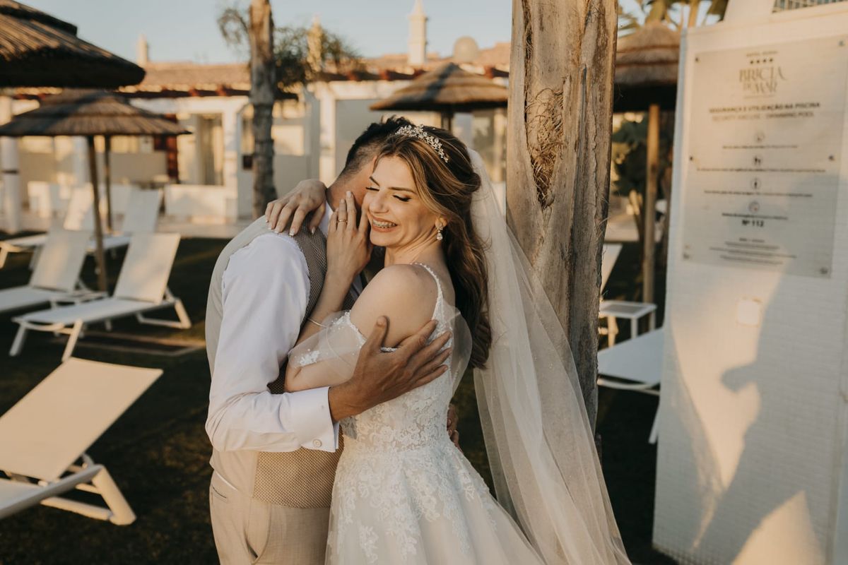 Bride in a lace gown and long veil resting her head against her groom, an Algarve village behind them