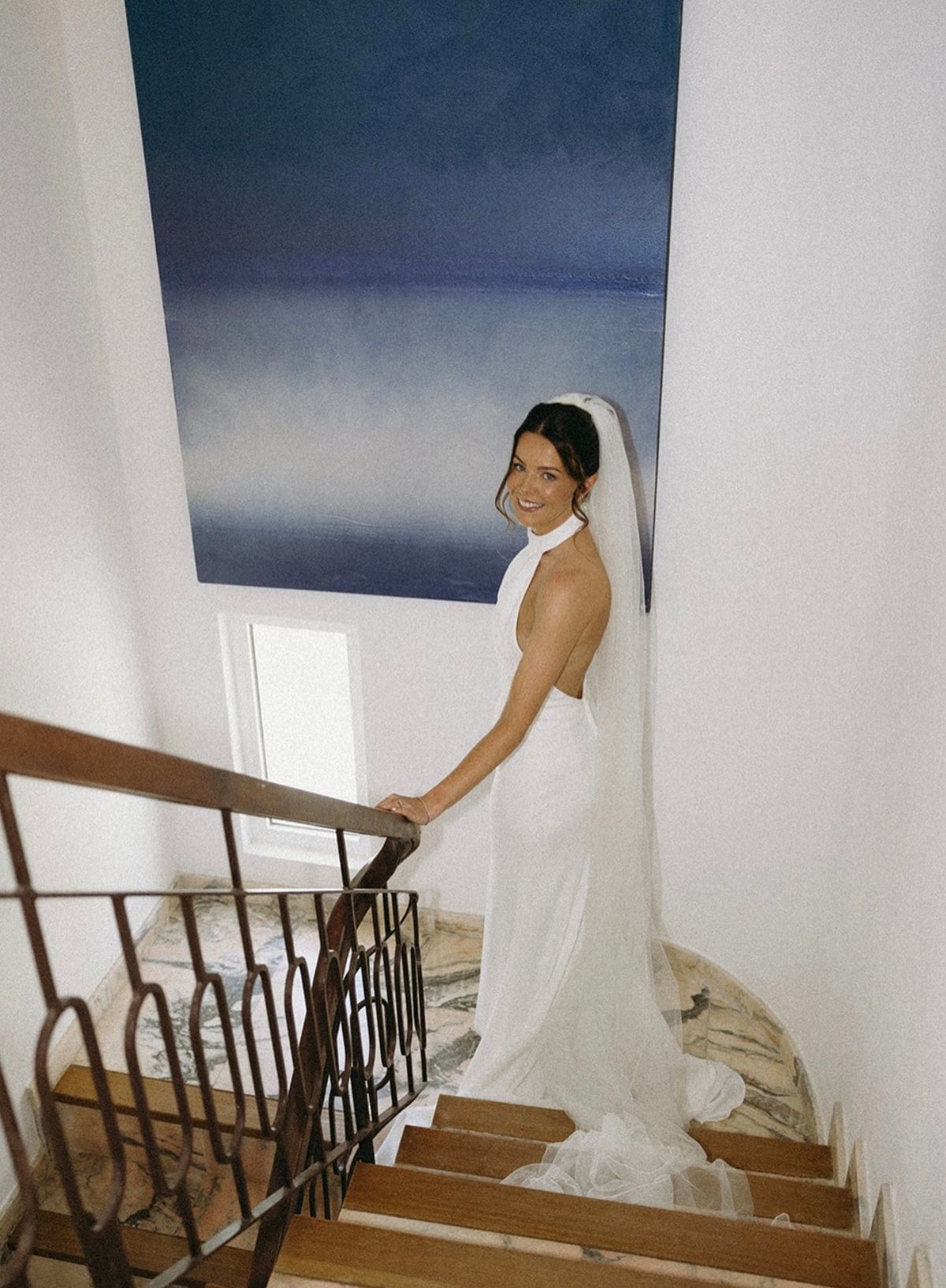 Bride in a fitted satin gown pausing on a staircase, her veil trailing behind her