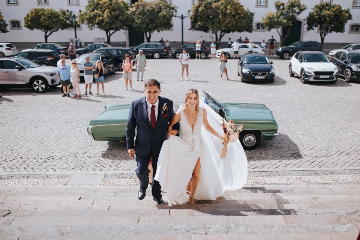 Bride arriving at the church on her father’s arm, crossing the village square past a vintage car