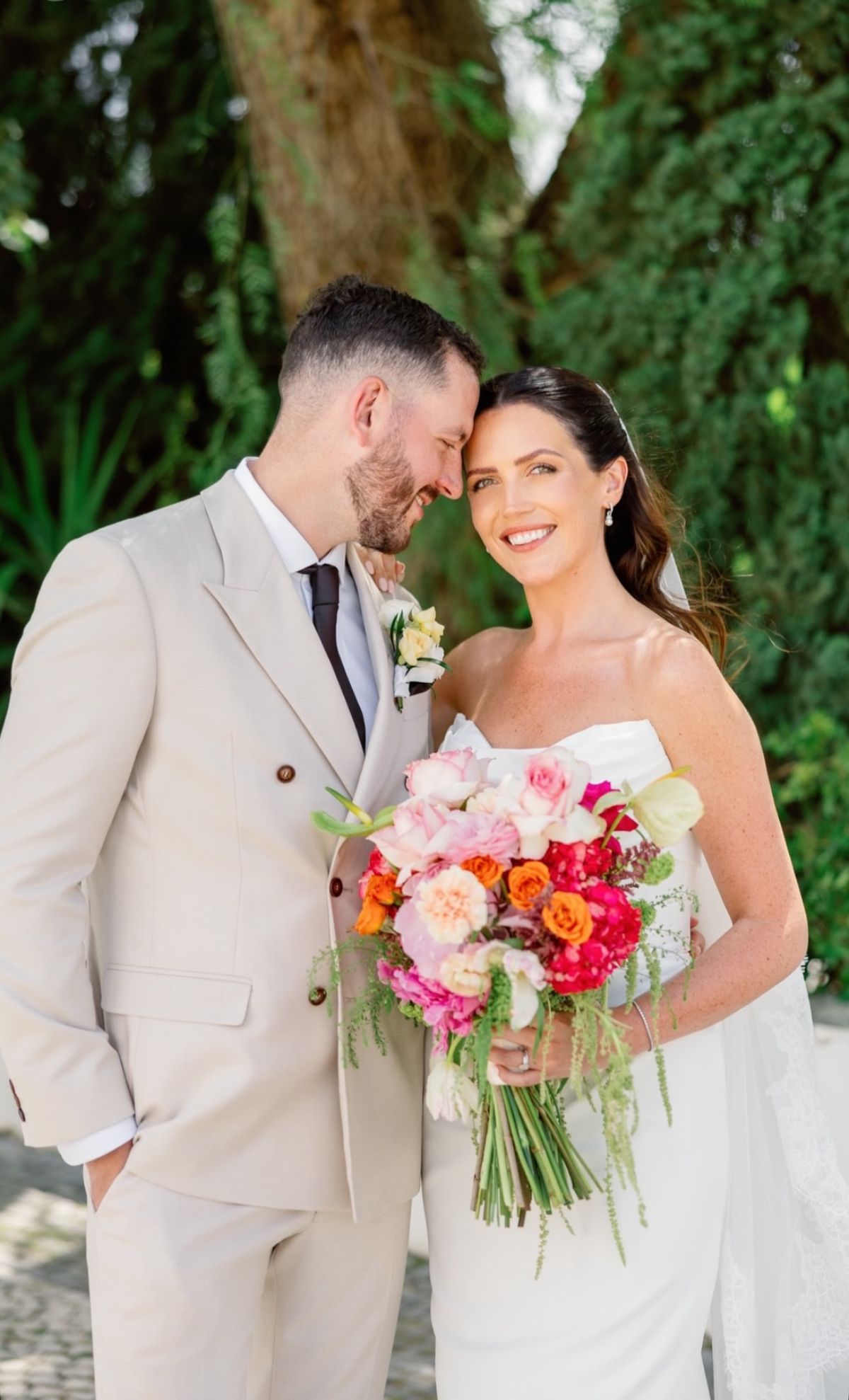 Bride and groom standing close together beneath a tree, the bride holding a bright coral and pink bouquet