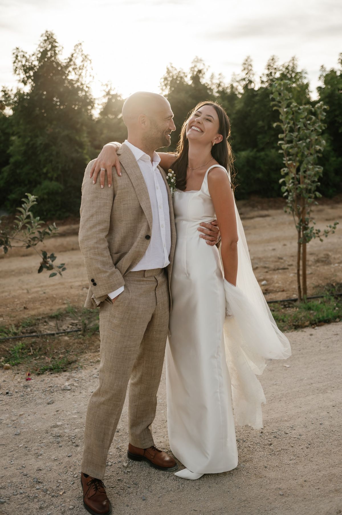Bride and groom walking together through an olive grove at golden hour, the bride in a simple satin gown