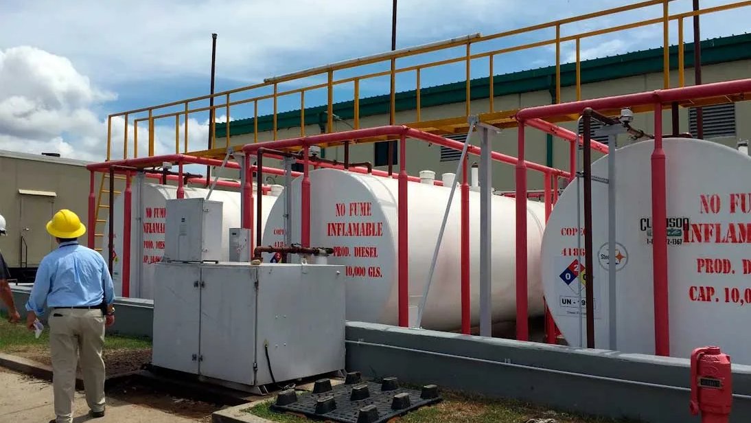 Técnico con casco amarillo inspecciona tanques horizontales blancos y sus tuberías rojas.