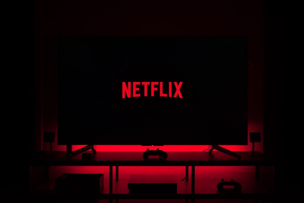 A dark room illuminated by the red glow of a television screen displaying the Netflix logo. The setting suggests a home entertainment environment, likely for watching movies or series. — Credit: Photo by Thibault Penin on Unsplash