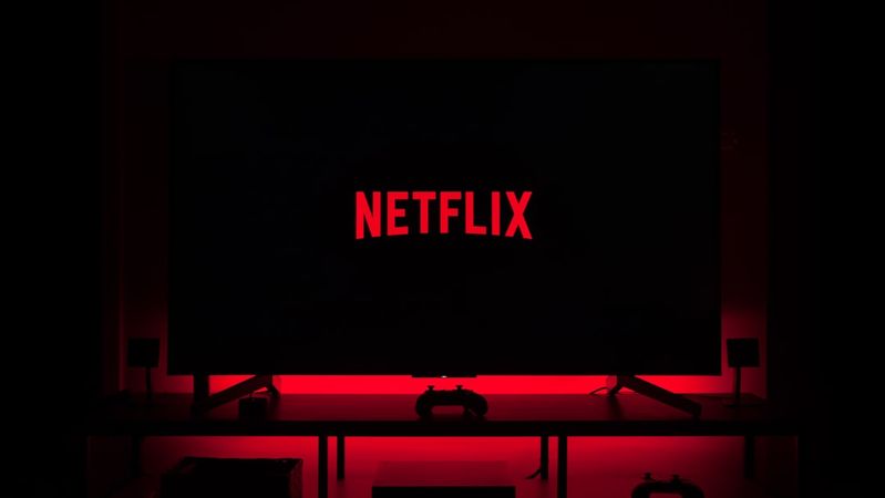 A dark room illuminated by the red glow of a television screen displaying the Netflix logo. The setting suggests a home entertainment environment, likely for watching movies or series.