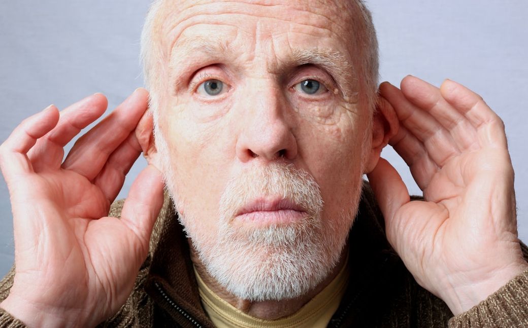 An elderly man is depicted placing his hands behind his ears, signaling a need to hear better. His expression suggests curiosity or attentiveness, creating a connection with the viewer. — Credit: Photo by Sharon Waldron on Unsplash