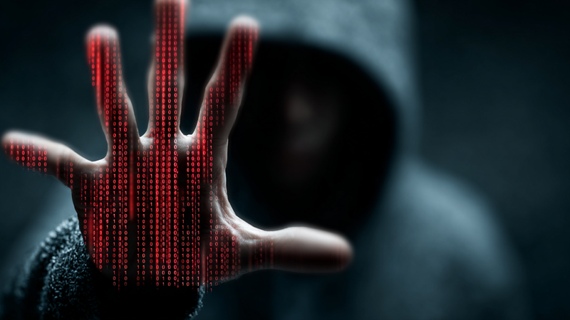 A figure in a dark hoodie reaches out with a hand, while streams of red binary code cascade down in front of them, creating a digital and ominous atmosphere. The blurred background suggests an environment focused on technology and cybersecurity.