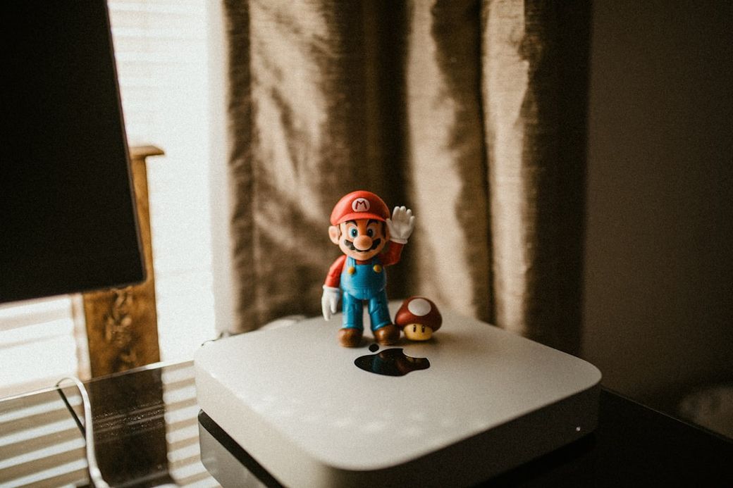 The image features a figurine of a popular video game character, Mario, standing on top of a white Apple device. Another small figurine, resembling a mushroom, is placed next to Mario, creating a playful and nostalgic scene. — Credit: Photo by Omar Rodriguez on Unsplash