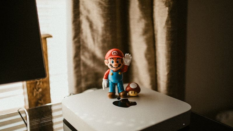 The image features a figurine of a popular video game character, Mario, standing on top of a white Apple device. Another small figurine, resembling a mushroom, is placed next to Mario, creating a playful and nostalgic scene.