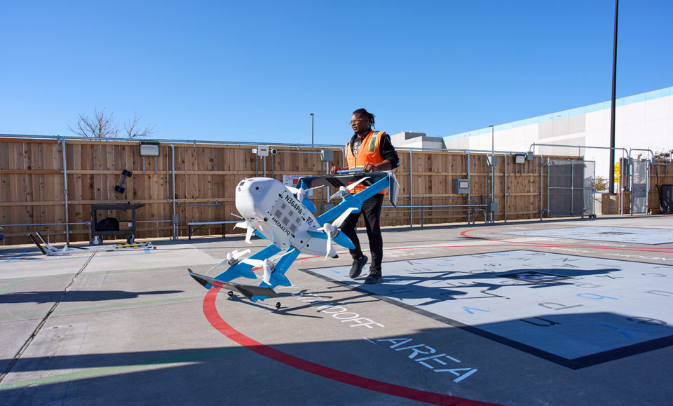 A person in an orange safety vest is transporting a drone on a helipad with markings indicating a takeoff area. The setting appears to be a facility designed for aviation or drone operations, with clear blue skies in the background.
