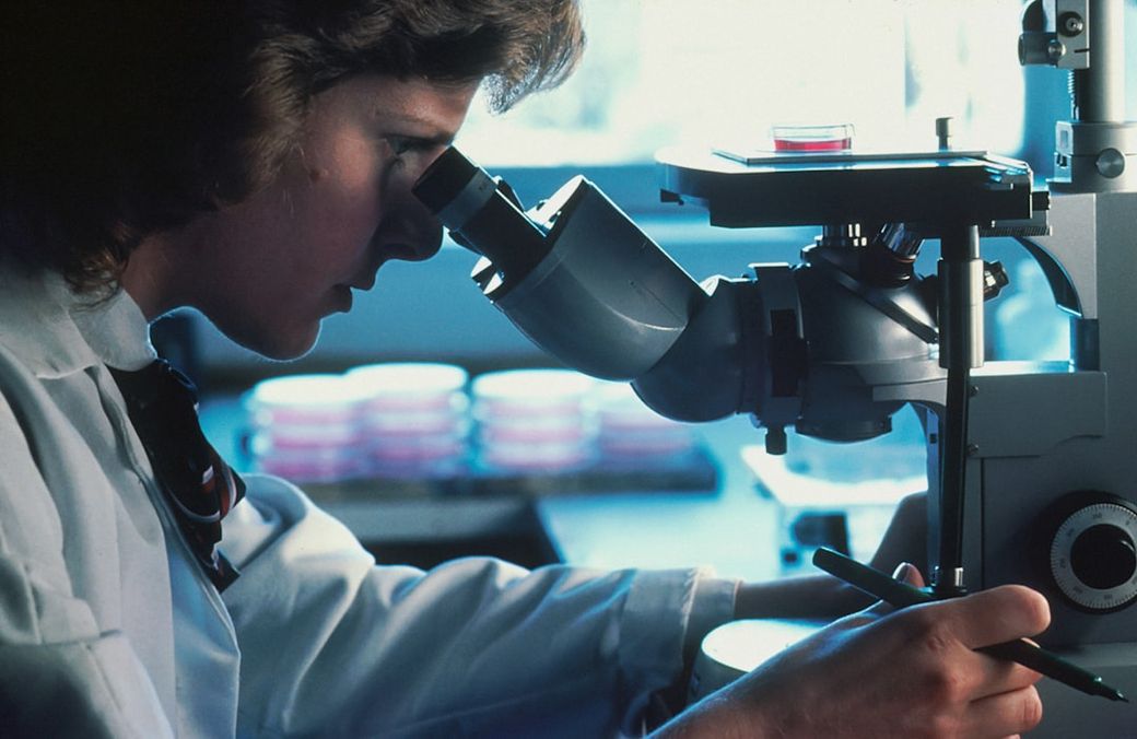 A scientist is using a microscope in a laboratory setting, focusing intently on her work. The background features petri dishes, indicating a biological or chemical research environment. — Credit: Photo by National Cancer Institute on Unsplash