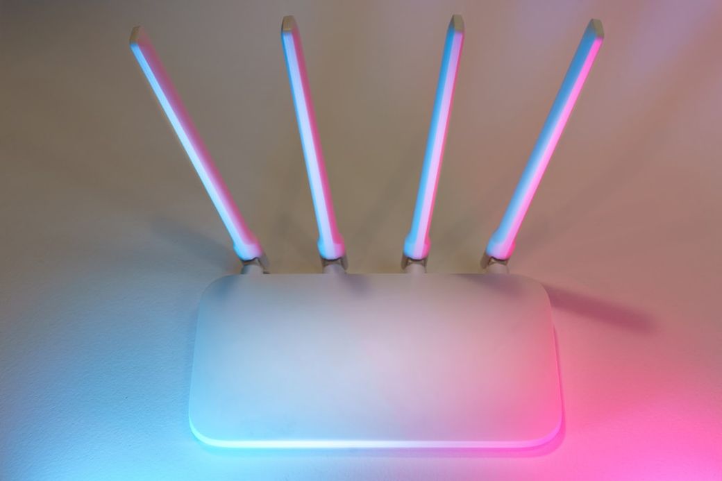 The image features a modern wireless router with four antennas, illuminated by colorful blue and pink lighting. The minimalist design emphasizes a sleek and contemporary aesthetic. — Credit: Photo by Jakub Żerdzicki on Unsplash