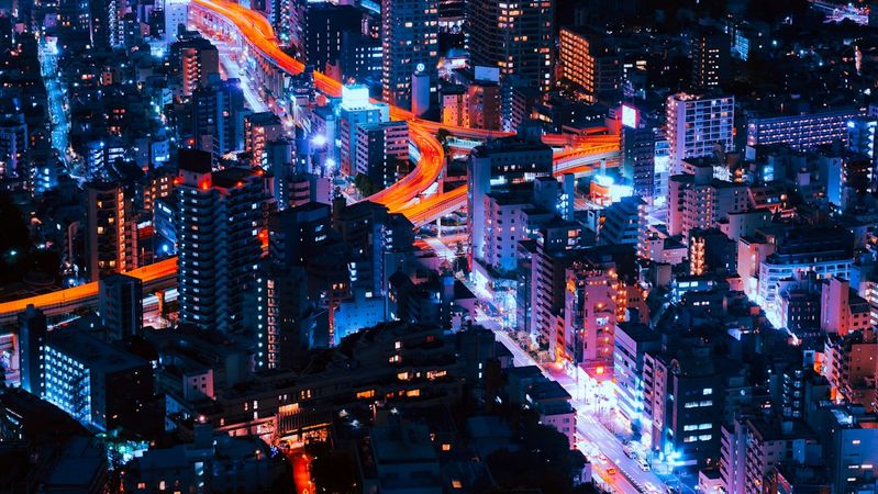 The image showcases a vibrant aerial view of a bustling cityscape at night, illuminated by a multitude of colorful lights. Skyscrapers and urban infrastructure are prominently featured, reflecting the dynamic energy of urban life.