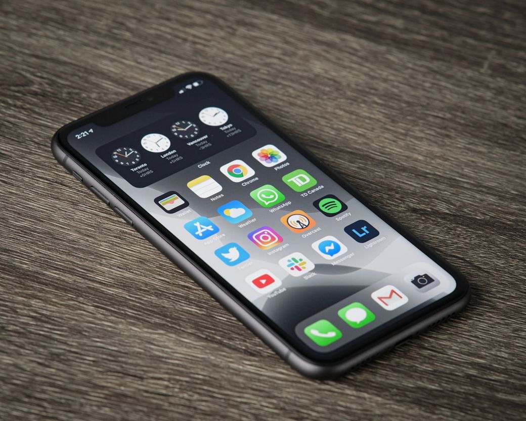 The image shows a close-up view of a smartphone lying on a textured surface. The screen displays various app icons and time widgets, indicating a modern digital device. — Credit: Photo by Thom Bradley on Unsplash