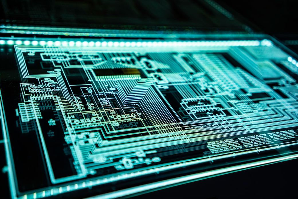 The image features a close-up view of a printed circuit board with intricate layered circuitry and connections. The design exhibits a glowing blue and green aesthetic, highlighting the complexity of electronic components and pathways. — Credit: Photo by Adi Goldstein on Unsplash