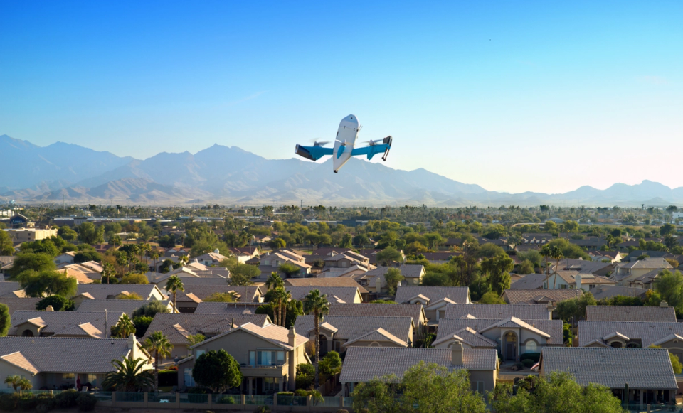 An aerial view of a residential neighborhood showcasing houses with light-colored roofs, beneath a clear blue sky. In the foreground, an advanced flying drone is seen ascending above the houses, with a mountainous backdrop.