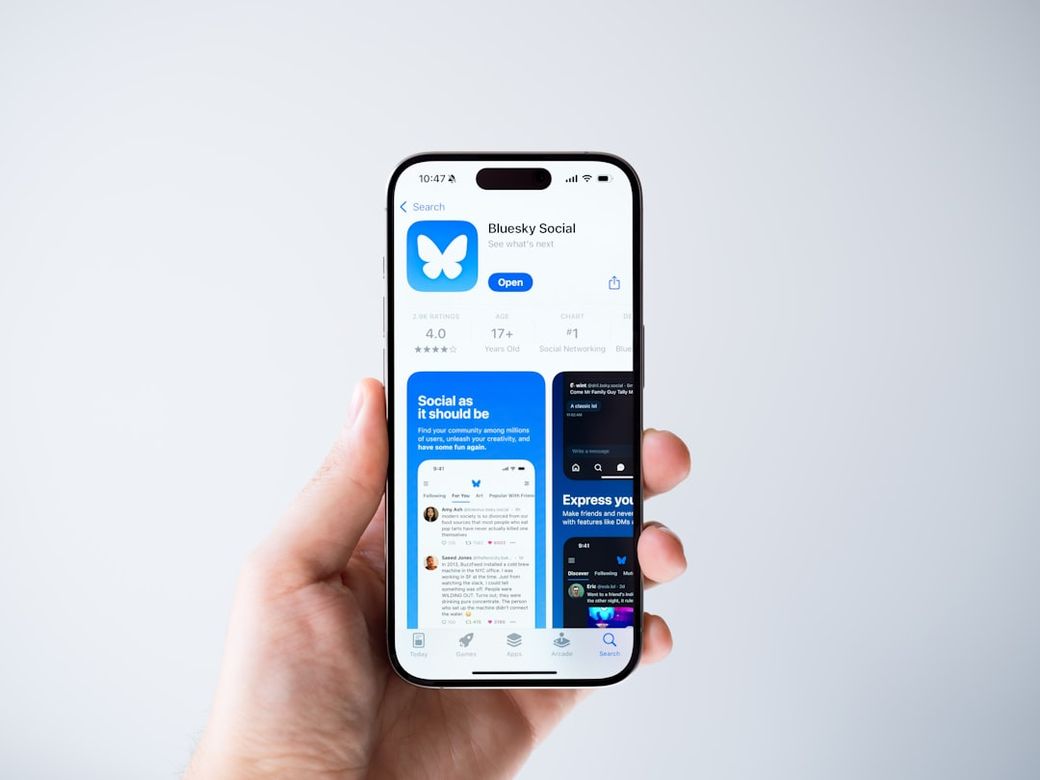 The image features a person holding a smartphone displaying the Bluesky Social app on its screen. The app showcases various social media features and user engagement elements. — Credit: Photo by Yohan Marion on Unsplash