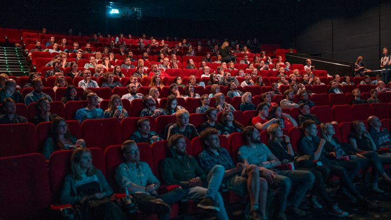 The image shows a full cinema auditorium filled with an audience watching a film. The red seating and dimly lit environment indicate that the movie is currently playing.