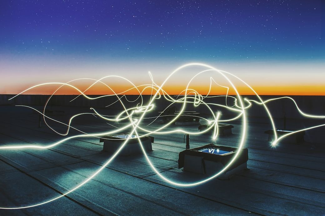 The image captures a rooftop scene at dusk, featuring abstract light trails against a colorful sky. The setting showcases a tranquil atmosphere with faint silhouettes of structures in the foreground. — Credit: Photo by Federico Beccari on Unsplash