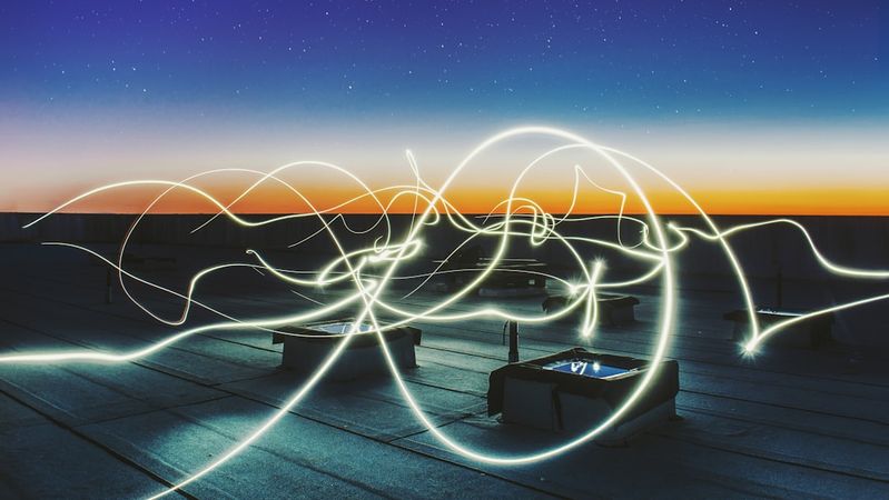 The image captures a rooftop scene at dusk, featuring abstract light trails against a colorful sky. The setting showcases a tranquil atmosphere with faint silhouettes of structures in the foreground.