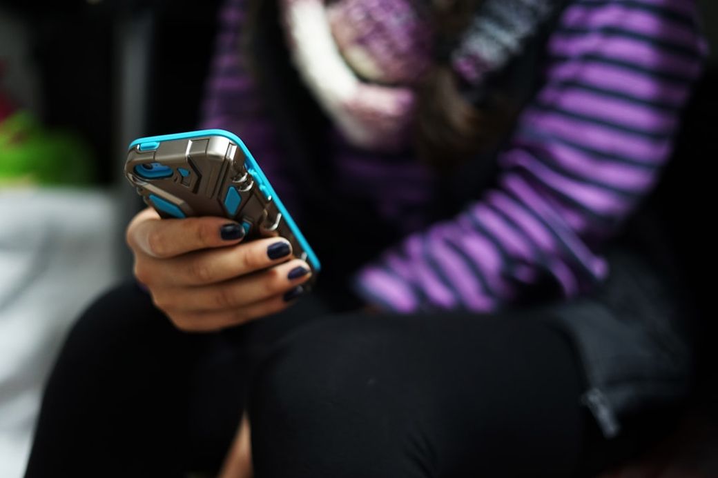 A close-up image of a person holding a smartphone, while seated. The individual is wearing a striped purple and black top and has their nails painted dark. — Credit: Photo by Julie Ricard on Unsplash