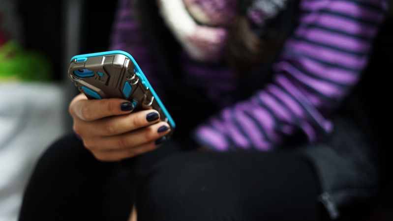 A close-up image of a person holding a smartphone, while seated. The individual is wearing a striped purple and black top and has their nails painted dark.