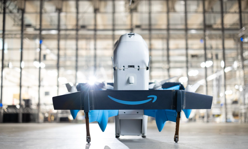 The image features an Amazon delivery drone positioned in a warehouse setting. The drone is shown from the front, highlighting its design and the prominent Amazon logo on its underside.