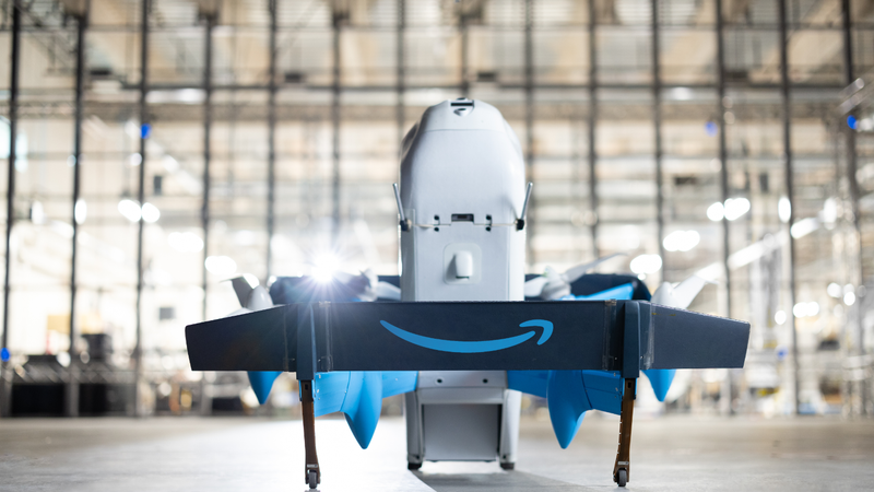 The image features an Amazon delivery drone positioned in a warehouse setting. The drone is shown from the front, highlighting its design and the prominent Amazon logo on its underside.
