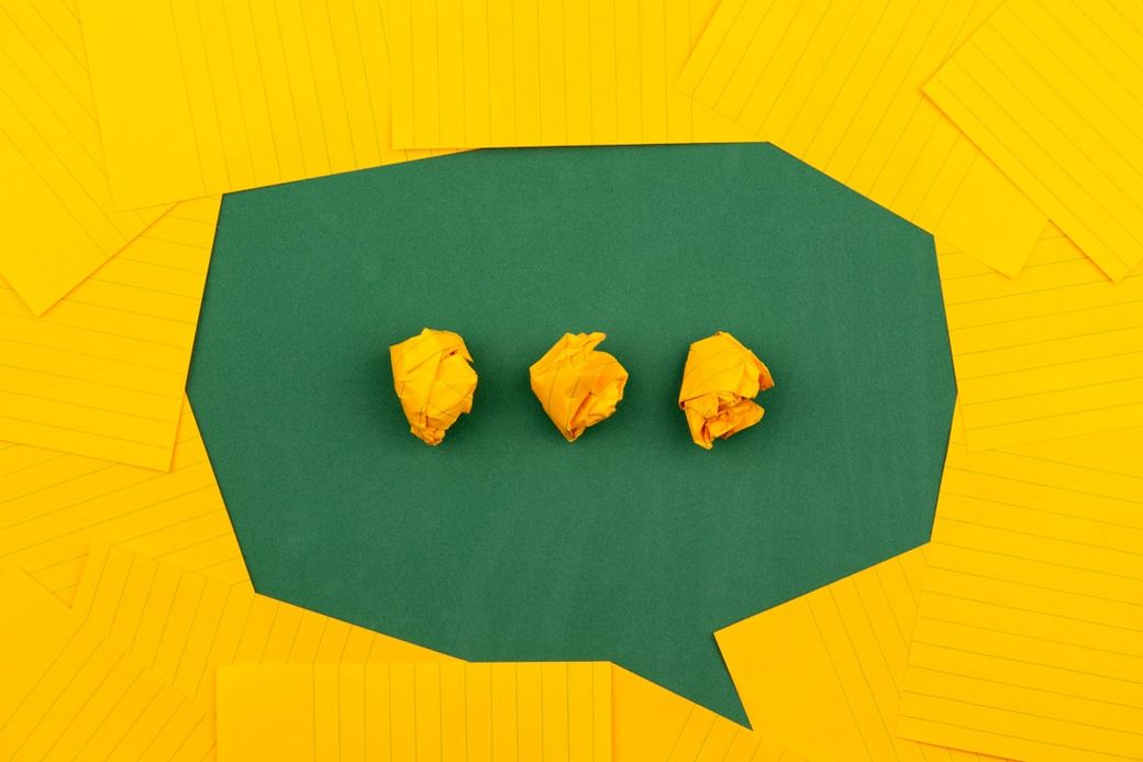The image features three crumpled yellow paper balls arranged on a green surface that is shaped like a speech bubble. Surrounding the bubble are scattered sheets of lined yellow paper, creating a vibrant and creative atmosphere. — Credit: Photo by Volodymyr Hryshchenko on Unsplash