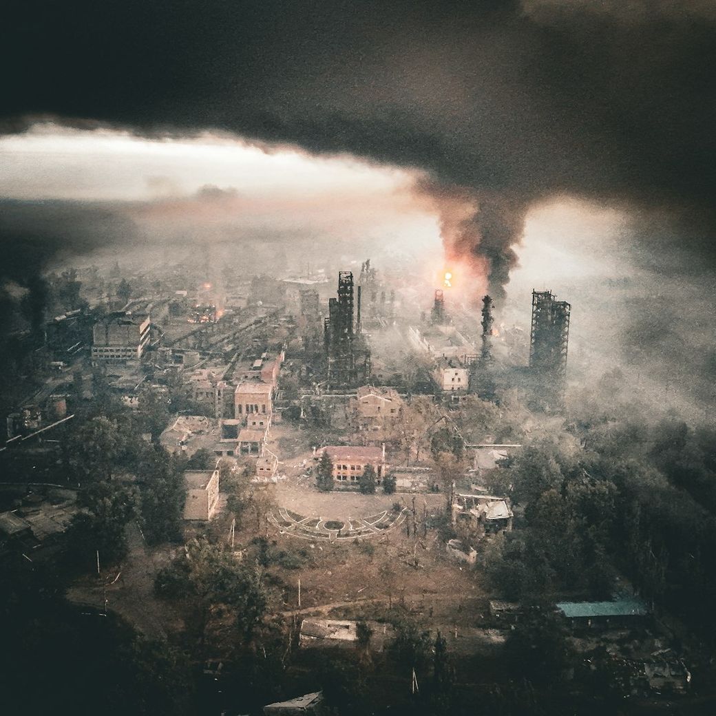 An aerial view of a devastated industrial area engulfed in smoke and flames. The scene depicts significant destruction with buildings partially collapsed and a large plume of smoke rising into the sky. — Credit: Photo by Serhii Kalyn on Unsplash