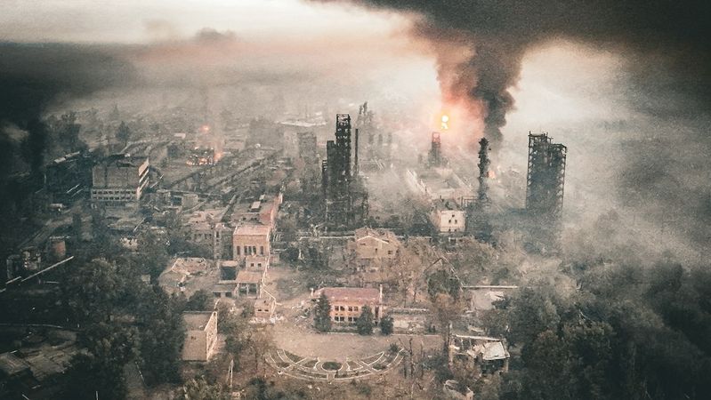 An aerial view of a devastated industrial area engulfed in smoke and flames. The scene depicts significant destruction with buildings partially collapsed and a large plume of smoke rising into the sky.