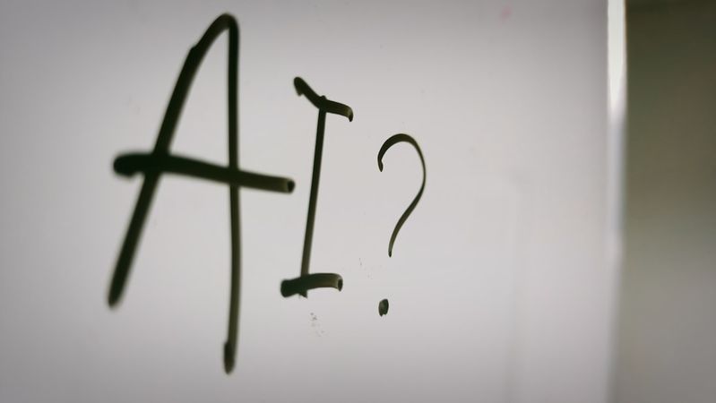 The image features the letters 'AI?' written on a surface, suggesting a contemplation or inquiry about artificial intelligence. The focus is on the text, which appears to be scrawled or drawn in a casual manner.