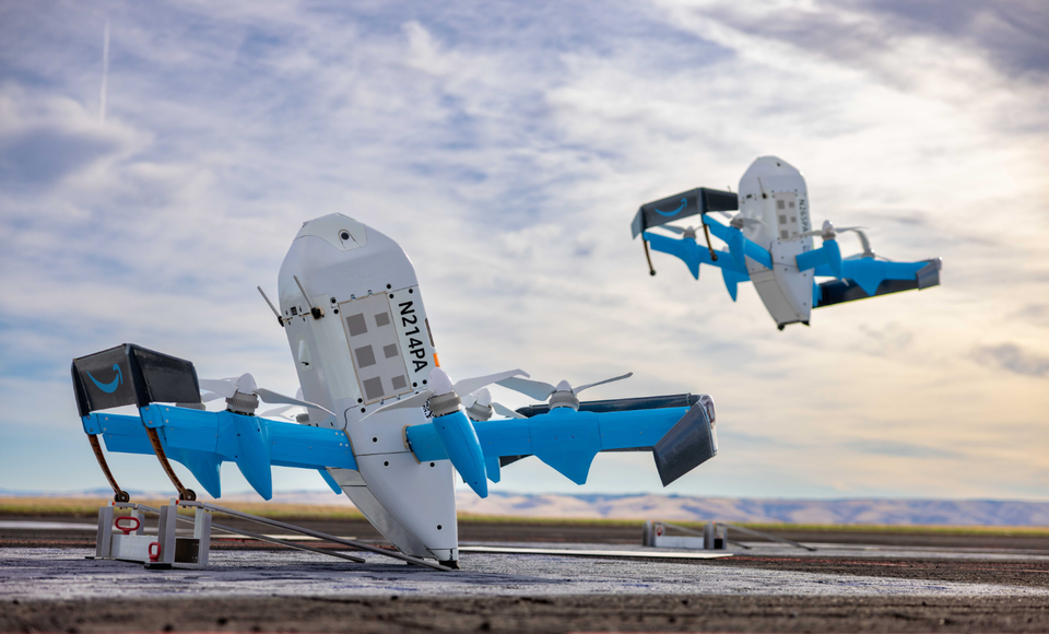 The image depicts two delivery drones taking off in a large outdoor space, with a cloudy sky in the background. The drones are equipped with blue and white designs, suggesting they are commercially branded.