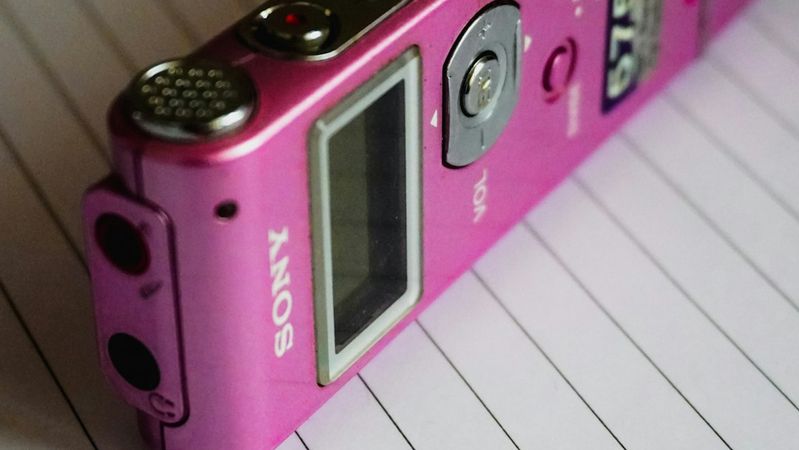 The image depicts a pink Sony voice recorder placed on lined notebook paper. The device features buttons on its side, indicating its functionality for recording audio.
