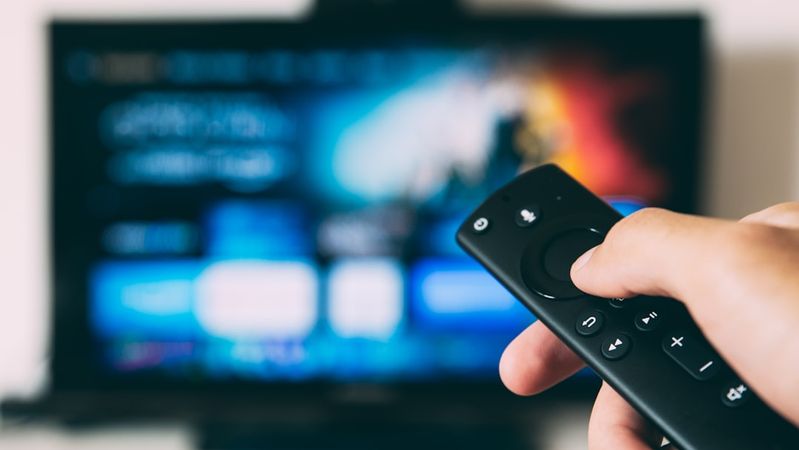 An individual is holding a remote control in front of a television displaying a colorful user interface. The screen features icons and options typical of a streaming service, suggesting a viewing experience.