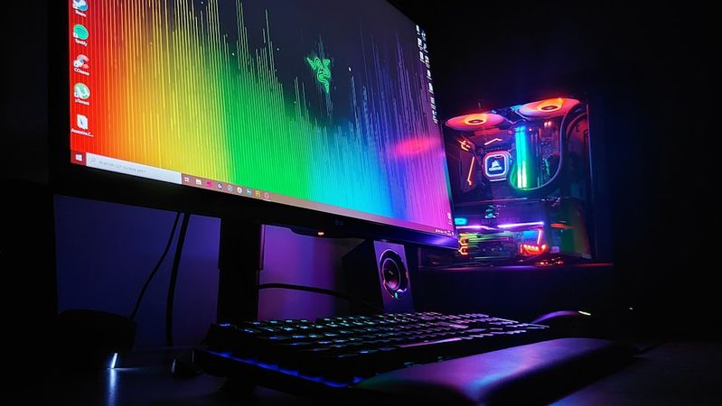The image features a dark room illuminated by the colorful glow of a gaming setup, including a monitor displaying a vibrant audio visualizer. A sleek computer tower with RGB lighting is visible, enhancing the ambient atmosphere.