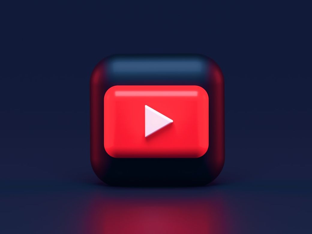 The image features a stylized YouTube play button icon set against a dark gradient background. The button is three-dimensional with rounded edges, emphasizing its visual appeal. — Credit: Photo by Alexander Shatov on Unsplash