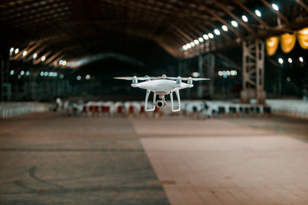 A drone is captured mid-flight in an expansive indoor space, with dim lighting and a row of empty chairs in the background. The scene conveys a sense of surveillance or high-tech innovation. — Credit: Photo by Adedotun Adegborioye on Unsplash