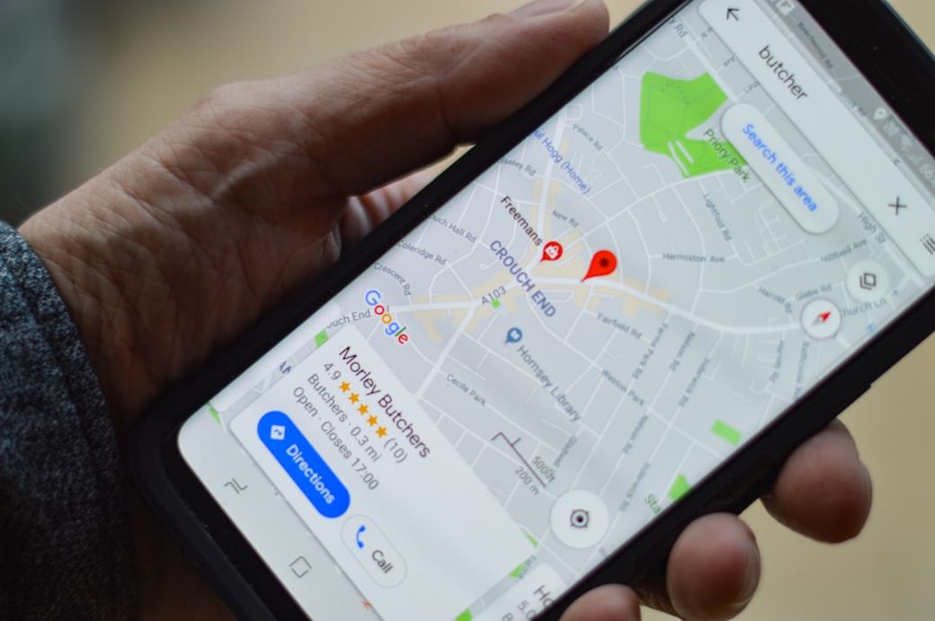 A close-up image of a hand holding a smartphone displaying a map application. The map shows a location marked with a red pin and various street names around it. — Credit: Photo by henry perks on Unsplash