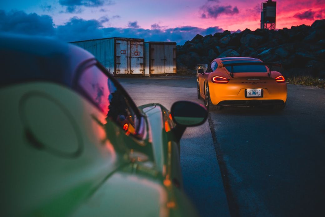 The image features two sports cars parked on a road with a dramatic sunset in the background. One car is brightly colored yellow, while the other is a contrasting green, indicating a vibrant automotive scene. — Credit: Photo by Erik Mclean on Unsplash