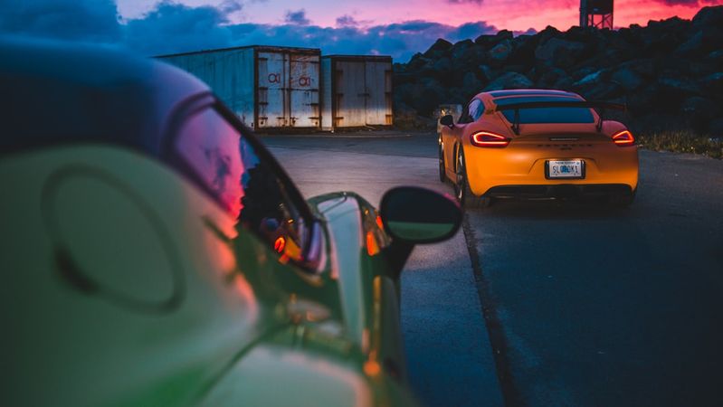 The image features two sports cars parked on a road with a dramatic sunset in the background. One car is brightly colored yellow, while the other is a contrasting green, indicating a vibrant automotive scene.