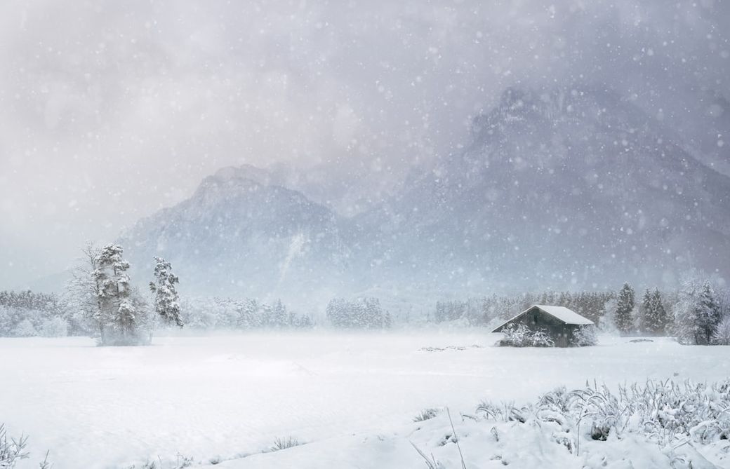 A serene winter landscape featuring a small cabin surrounded by a blanket of snow and tall mountains in the background. Snow is falling gently, creating a tranquil and peaceful atmosphere. — Credit: Photo by simon on Unsplash