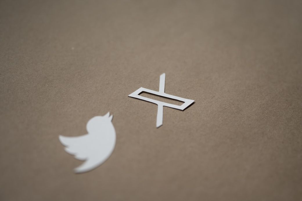 The image features two logos: the classic Twitter bird logo and a stylized 'X' logo. They are set against a plain brown background, highlighting their distinct shapes. — Credit: Photo by Kelly Sikkema on Unsplash