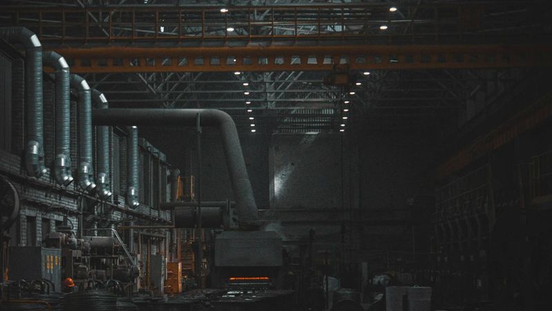 An industrial warehouse interior featuring a large, dimly lit space with high ceilings and structural beams. The scene includes machinery and production equipment, emphasizing the manufacturing aspect of the environment.